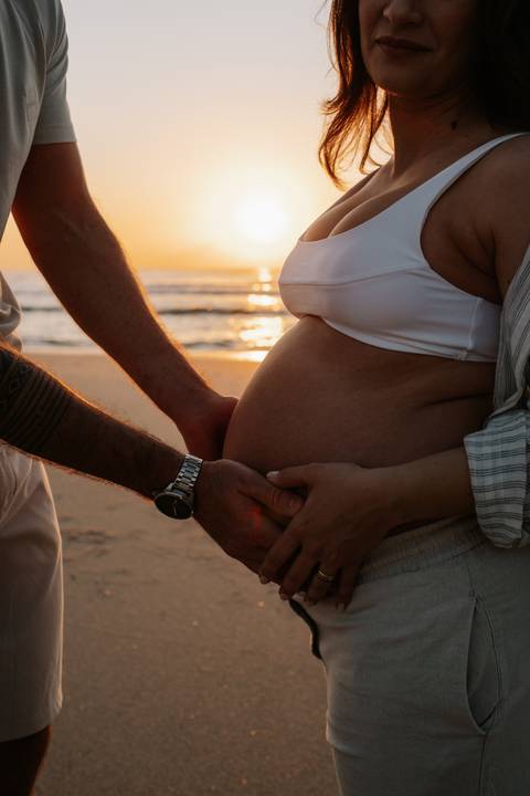 Foto em detalhe das mãos do casal formando um coração sobre a barriga na luz do sol.'
