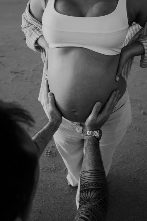 Foto em preto e branco do detalhe das mãos do pai sobre a barriga de grávida.'