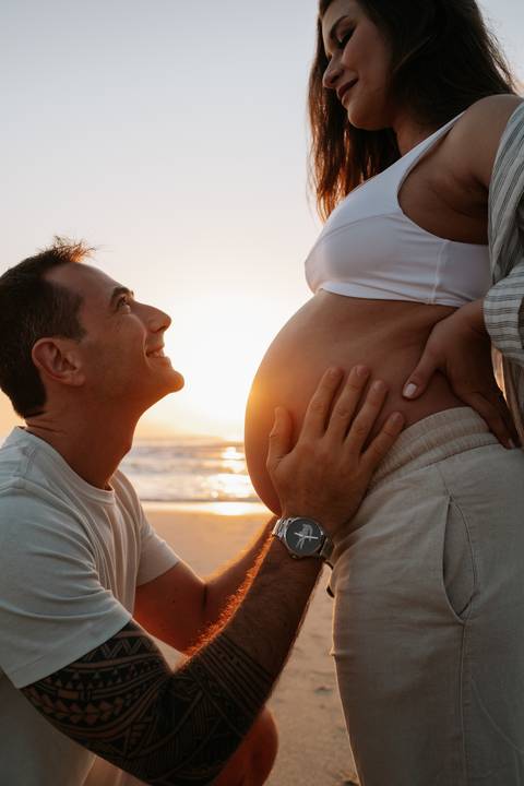 Foto do pai sorrindo e olhando para a gestante contra a luz do sol na praia.'