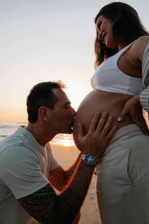 Foto do pai beijando a barriga da gestante sob a luz dourada do amanhecer.'