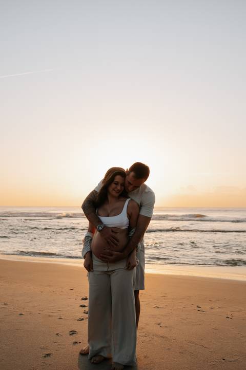 Foto do pai abraçando a gestante por trás em momento de silhueta no pôr do sol.'