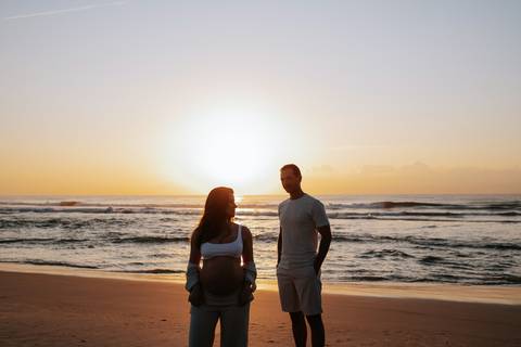 Foto da silhueta do casal frente a frente na beira do mar com o sol ao centro.'