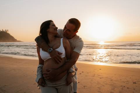 Foto do casal abraçado na areia com o mar e o sol no horizonte.'