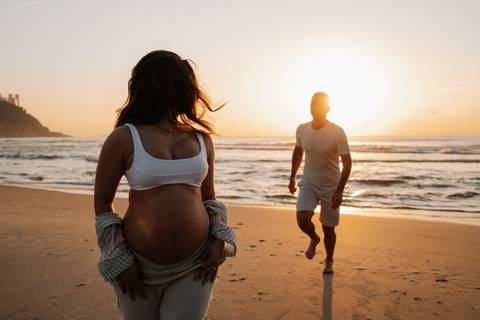 Foto da gestante em primeiro plano com o parceiro caminhando ao fundo na praia.'