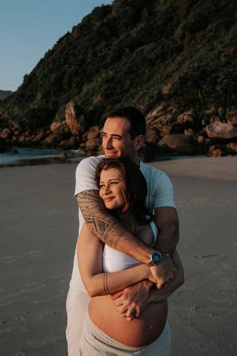 Foto do casal abraçado na areia com as rochas e a natureza ao fundo.'