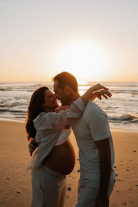 Foto do casal sorrindo e se abraçando com a luz do sol refletida na água.'