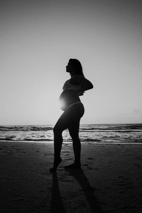 Foto em preto e branco da silhueta artística da gestante na beira do mar.'