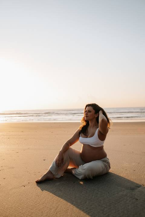 Foto da gestante sentada na areia contemplando o mar no nascer do sol.'
