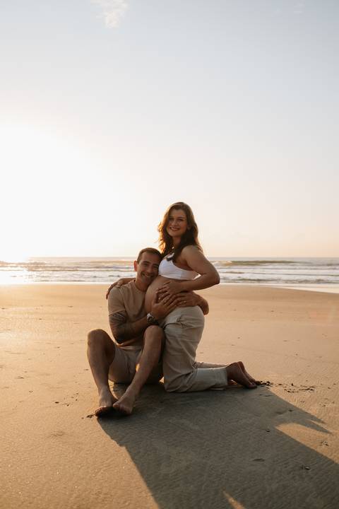 Foto do casal sorrindo sentado na areia com a luz dourada do sol.'