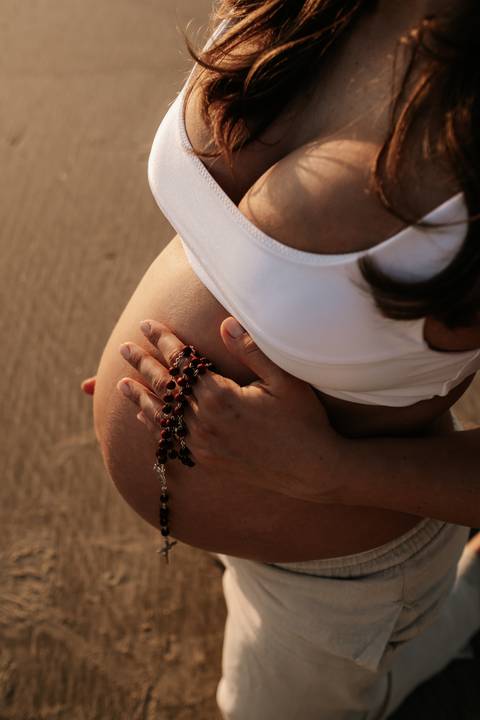 Foto em detalhe da mão da gestante segurando um terço sobre a barriga.'