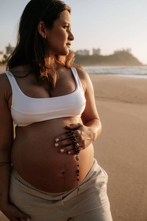Foto aproximada do terço sobre a barriga de grávida em momento de fé.'