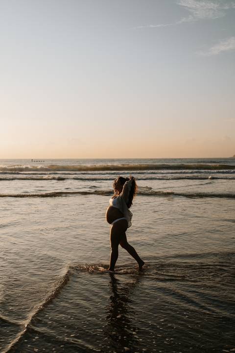 Foto da silhueta da gestante em pé na beira do mar com braços erguidos.'