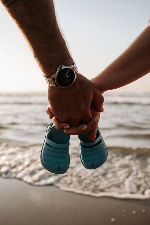 Foto em detalhe das mãos do casal segurando sapatinhos de bebê com o mar ao fundo.'