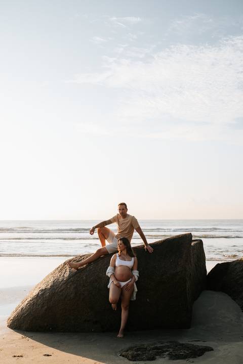Foto do pai sentado na rocha com a gestante à frente na beira do mar.'