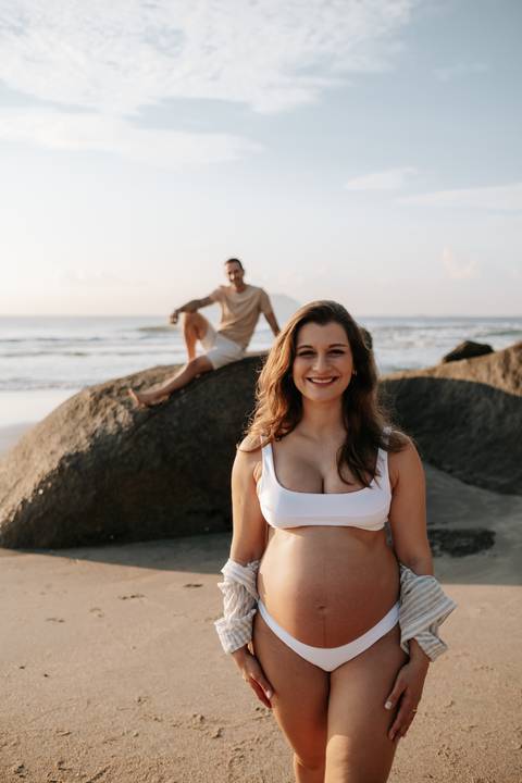 Foto aproximada da gestante sorrindo com o parceiro ao fundo nas pedras.'