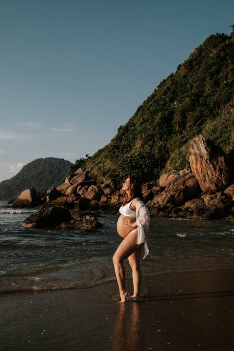 Foto de perfil da gestante em frente às rochas e vegetação da praia.'