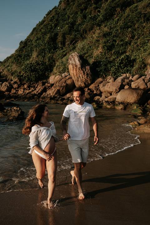 Foto do casal caminhando na beira do mar com rochas ao fundo.'
