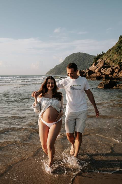 Foto da gestante de camisa branca com o parceiro na praia.'