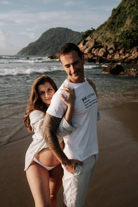 Foto da gestante abraçando o parceiro por trás na areia.'