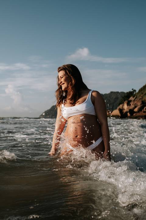 Foto da gestante sorrindo com as ondas quebrando ao fundo.'