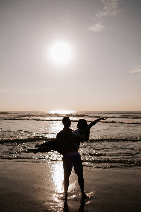 Foto da silhueta do pai erguendo a gestante no colo no sol.'