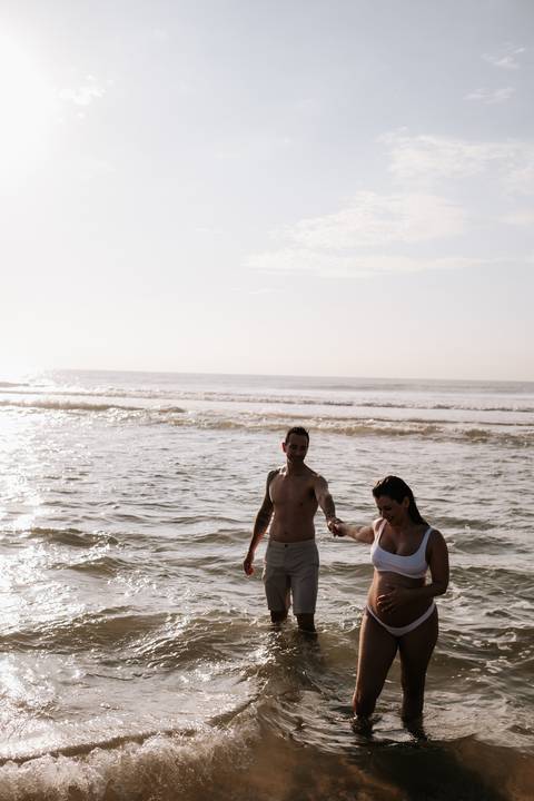 Foto do casal de mãos dadas caminhando na água em plano aberto.'