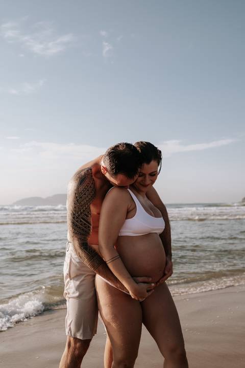 Foto aproximada do pai abraçando a gestante por trás com o mar ao fundo.'