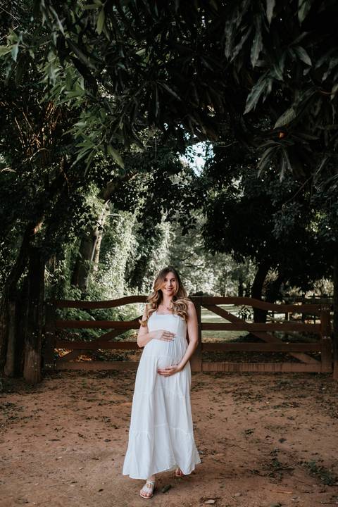 Foto da gestante de vestido branco em frente a uma porteira de madeira.'