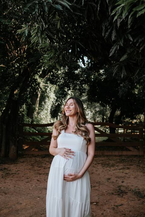 Foto da gestante de olhos fechados apreciando a luz entre as árvores.'