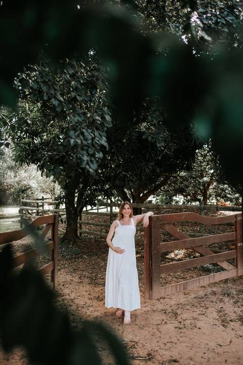 Foto da gestante posando ao lado da porteira com árvores ao fundo.'