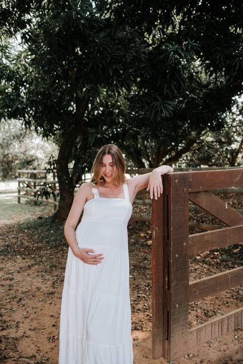 Foto da gestante encostada na cerca de madeira destacando a barriga.'