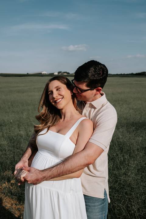 Foto aproximada do casal com o pai abraçando a gestante por trás.'
