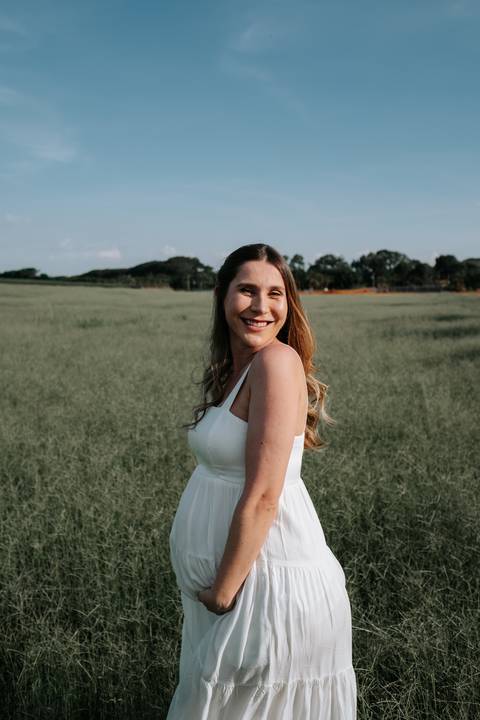 Foto da gestante de vestido branco sorrindo em plano médio.'