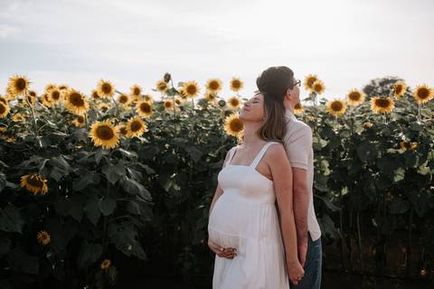 Foto do pai abraçando a gestante por trás no campo de flores.'