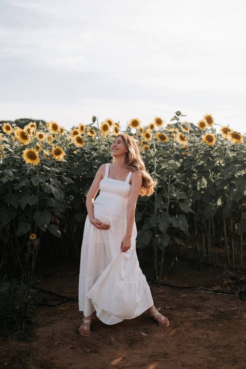 Foto da gestante em pé destacando a barriga no campo ensolarado.'