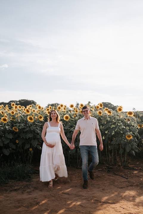 Foto do casal de mãos dadas em plano aberto entre girassóis.'