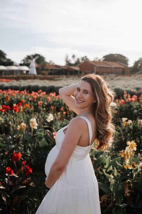 Foto de perfil da gestante sorridente em um campo de flores coloridas.'