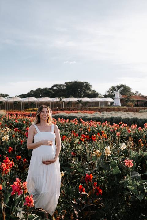 Foto da gestante posando em meio a flores vermelhas e brancas.'