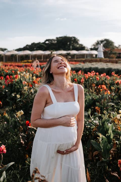 Foto da gestante rindo com as mãos na barriga no jardim.'
