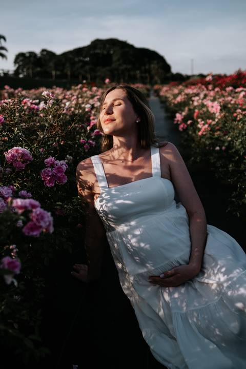 Foto de perfil da gestante sentada no jardim de flores.'