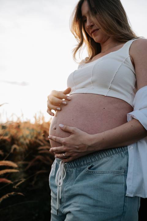 Foto em detalhe das mãos da gestante sobre a barriga exposta.'
