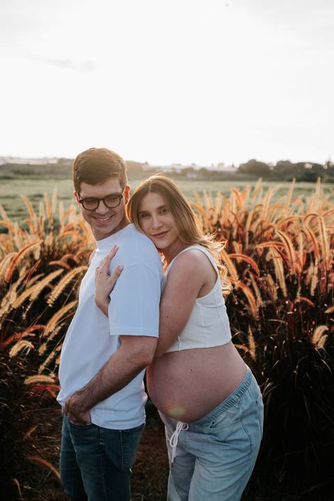Foto da gestante abraçando o parceiro por trás no campo.'