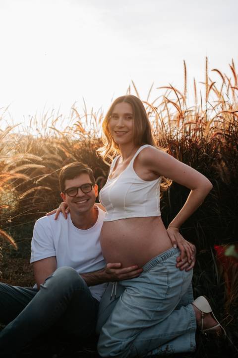 Foto do casal sorrindo entre capins altos no entardecer.'