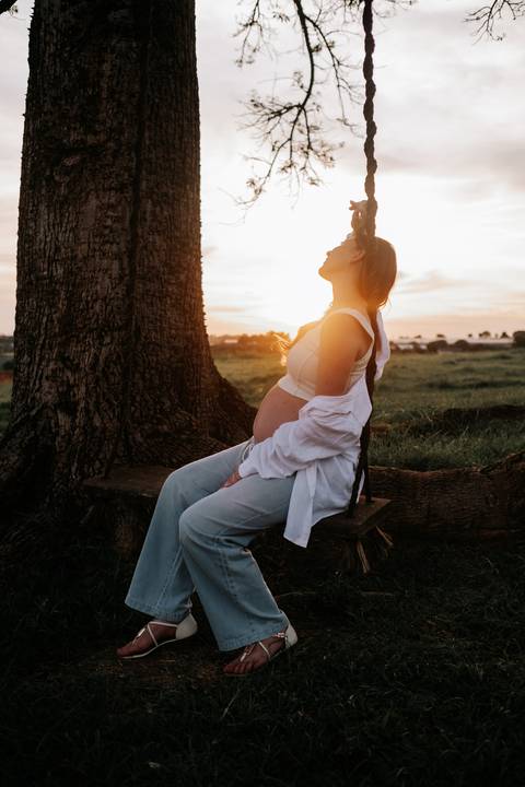 Foto da gestante sentada no balanço de madeira sob uma árvore.'