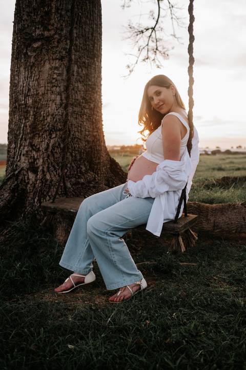 Foto da gestante sorrindo no balanço com camisa branca fluida.'