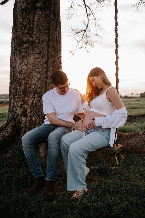 Foto do casal sentado no balanço interagindo com a barriga.'