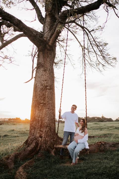 Foto do pai ao lado da gestante no balanço sob a árvore.'