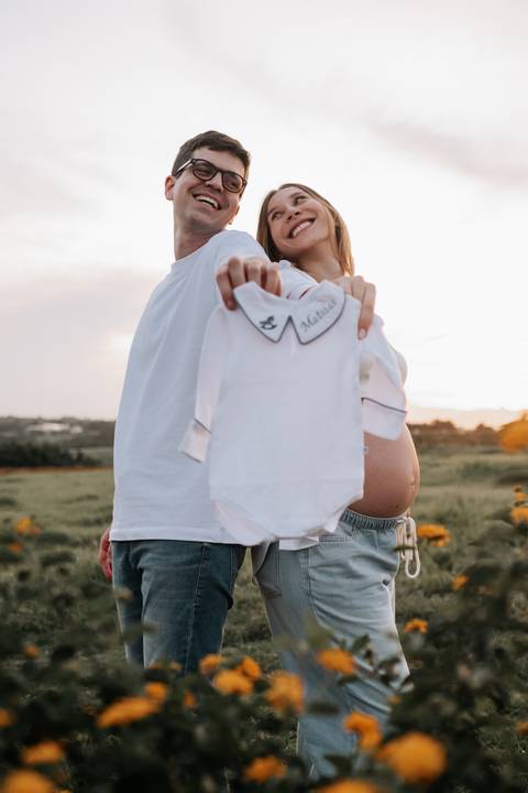 Foto do casal de costas segurando a roupinha do bebê.'