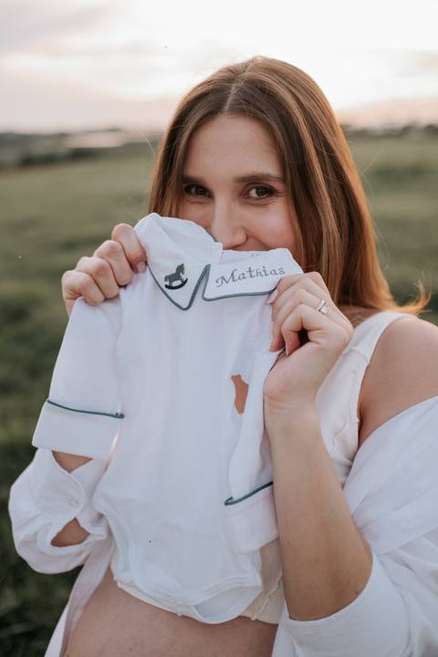 Foto da gestante sorrindo atrás da roupinha do Mathias.'