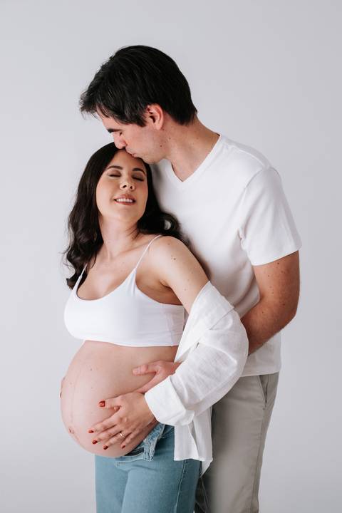 Foto do casal abraçado com o pai beijando o topo da cabeça da gestante.'